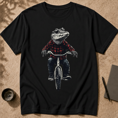A black short-sleeve crewneck t-shirt laid flat on a textured tan surface. The shirt features a graphic print of an alligator wearing a red and black plaid flannel shirt and riding a bicycle.