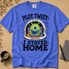 A royal blue graphic t-shirt laid flat on a textured tan surface, featuring a cartoon green one-eyed alien reading in a space helmet with the text 'PLOT TWIST: I STAYED HOME'. A coffee cup, pen, and notebook are styled around the shirt.
