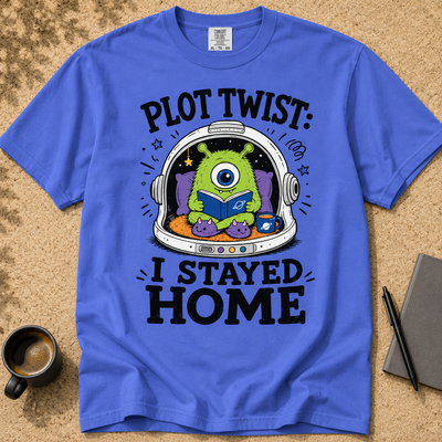 A royal blue graphic t-shirt laid flat on a textured tan surface, featuring a cartoon green one-eyed alien reading in a space helmet with the text 'PLOT TWIST: I STAYED HOME'. A coffee cup, pen, and notebook are styled around the shirt.
