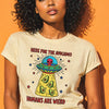 Woman with voluminous curly black hair wearing cream t-shirt featuring "Here For The Avocados" alien UFO beaming avocados graphic with "Humans Are Weird" text