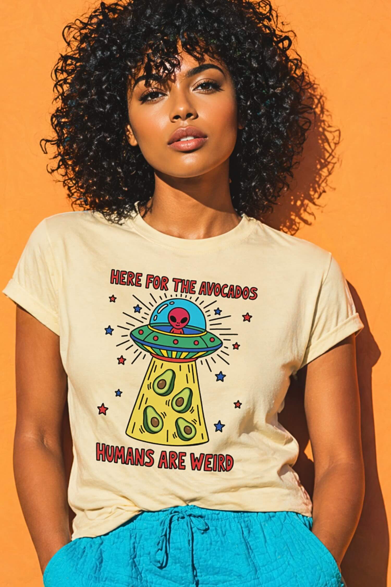 Woman with voluminous curly black hair wearing cream t-shirt featuring "Here For The Avocados" alien UFO beaming avocados graphic with "Humans Are Weird" text